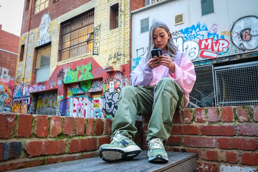 A photo of an Asian girl sitting on a brick wall using her mobile with graffiti behind her.