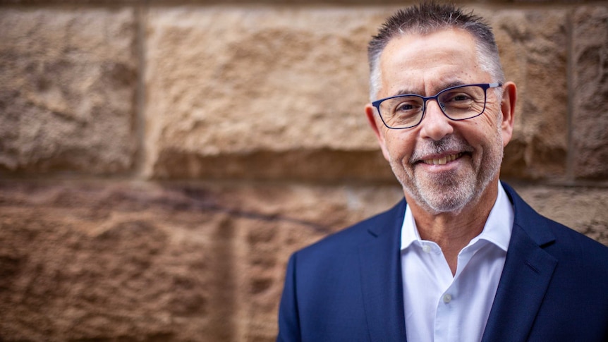 Norman Swan smiles in front of a brick wall. He wears a navy blazer and a crisp white shirt.