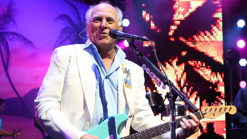 Jimmy Buffett performs in a white jacket playing a blue guitar.