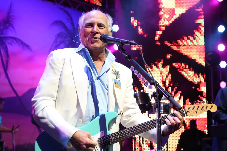 Jimmy Buffett performs in a white jacket playing a blue guitar.