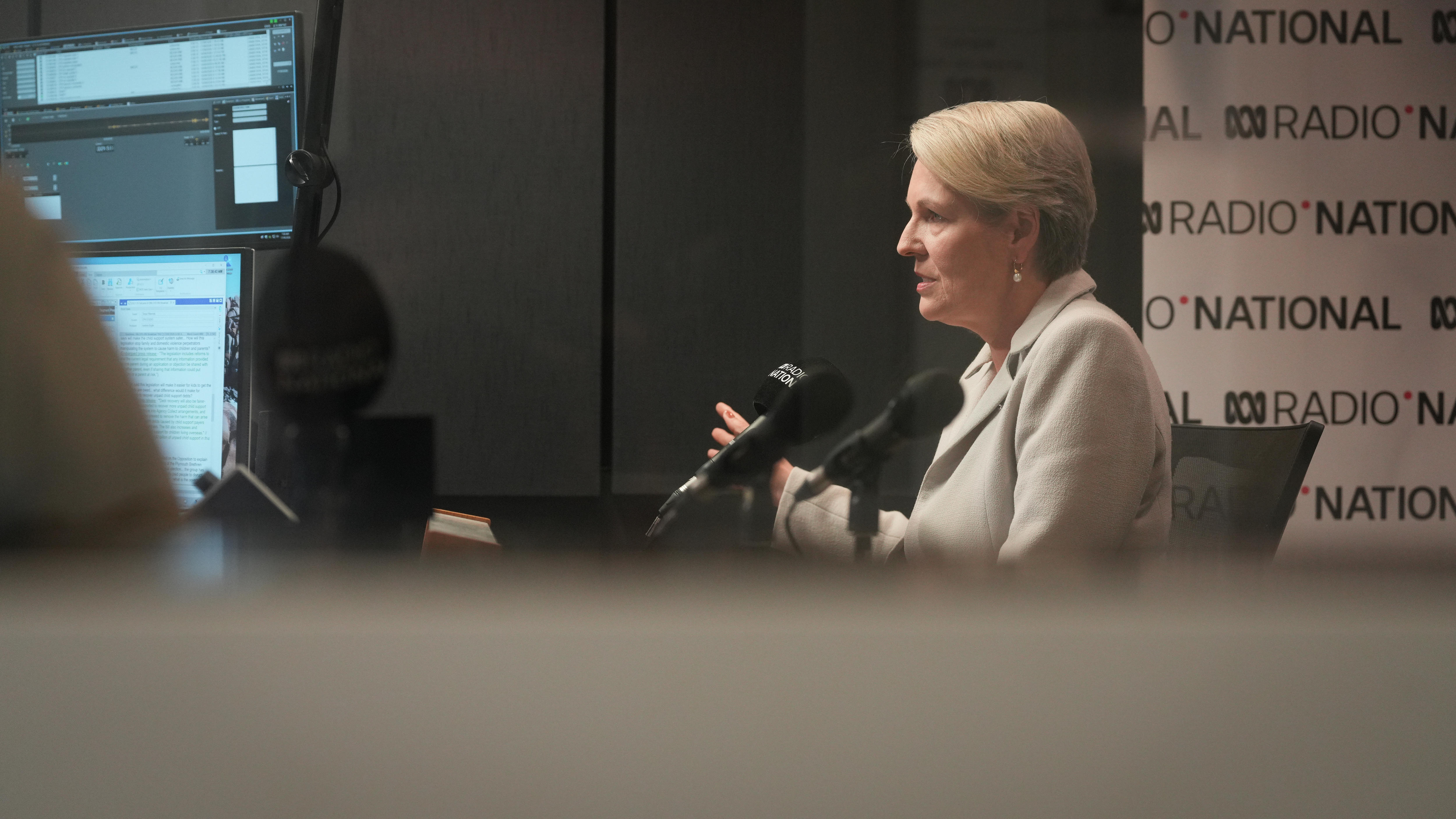 A woman in a grey suit speaking in a radio studio with a Radio National banner behind her
