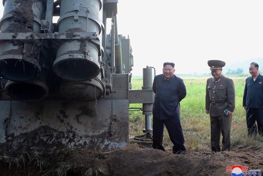 North Korean leader Kim Jong-un stands next to large cylinder structure in the dirt.