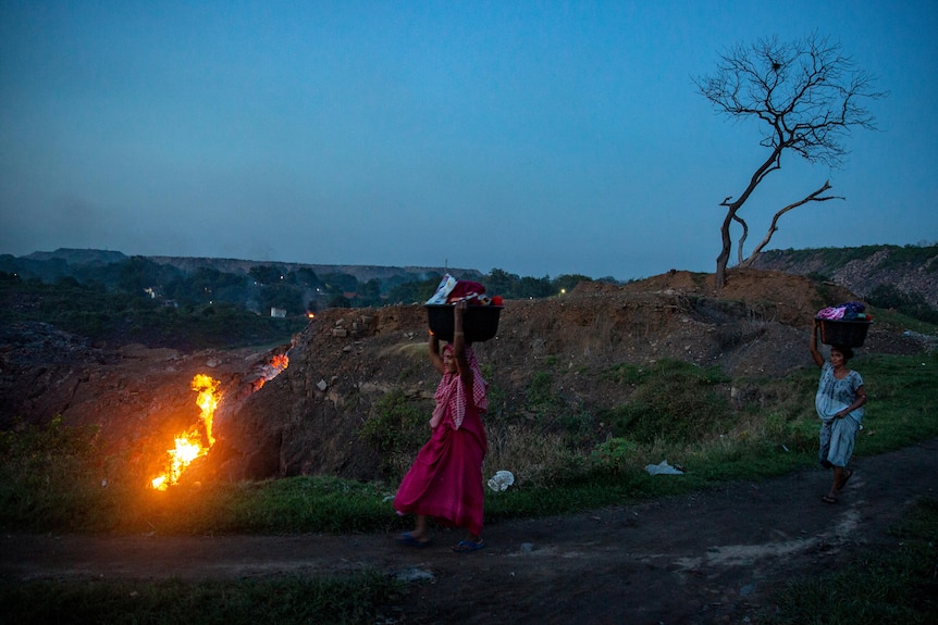 Indian women walk past flickering coal mine at dusk
