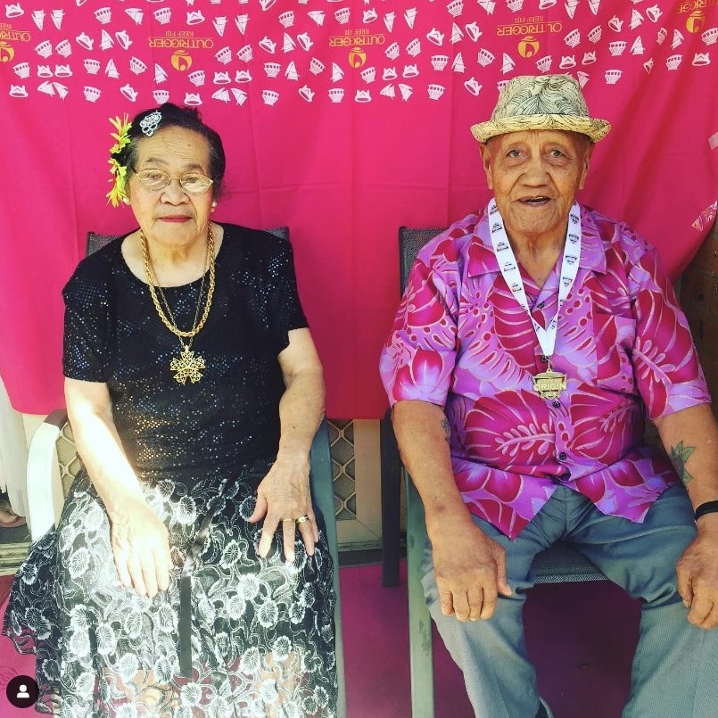An elderly Tongan woman with flowers in her hair and a Tongan man in a bright purple shirt smiling
