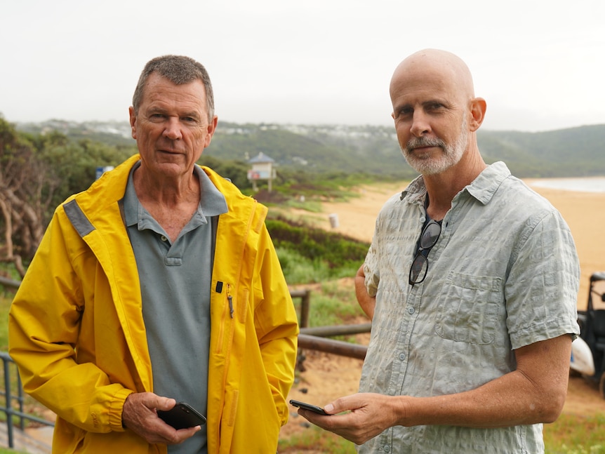 A man in a yellow raincoat standing next to a man in a tshirt both holding phones at the beach.