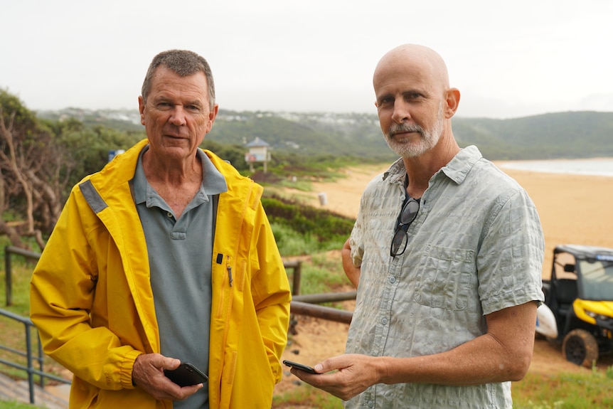 A man in a yellow raincoat standing next to a man in a tshirt both holding phones at the beach.
