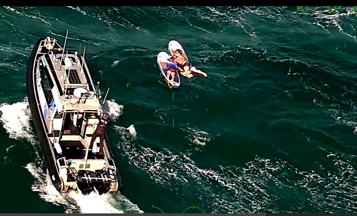 A police boat near two people lying on paddleboards in a green choppy sea.