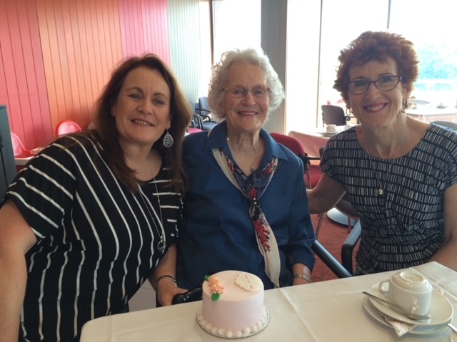 Three women sit next to each other in a cafe, leaning together and smiling.