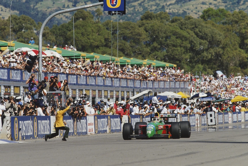 A man in a yellow jacket waves the chequered flag as a Formula 1 car speeds past and a sea of faces watch from the grandstand.