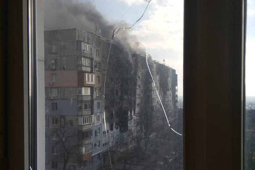 Black smoke plumes from a building seen through cracked glass.