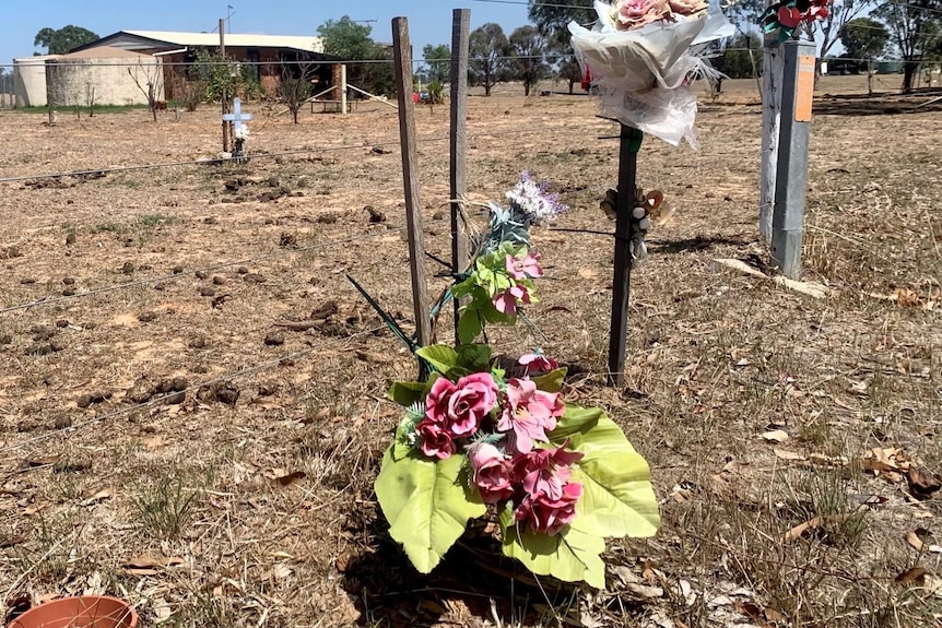 Three bunches of flowers tied to a wire fence with a memorial cross further off in the field.