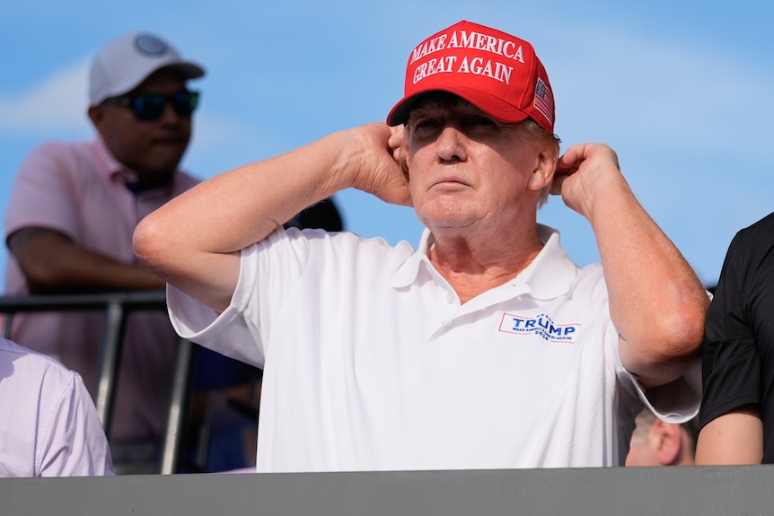 A man in a white shirt adjusts his red hat that says "make American great again"