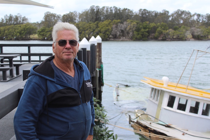 An elderly man, Karl Krieter, standing with his hands in his pockets on a boat ramp. In front of a sunken vessel in the water