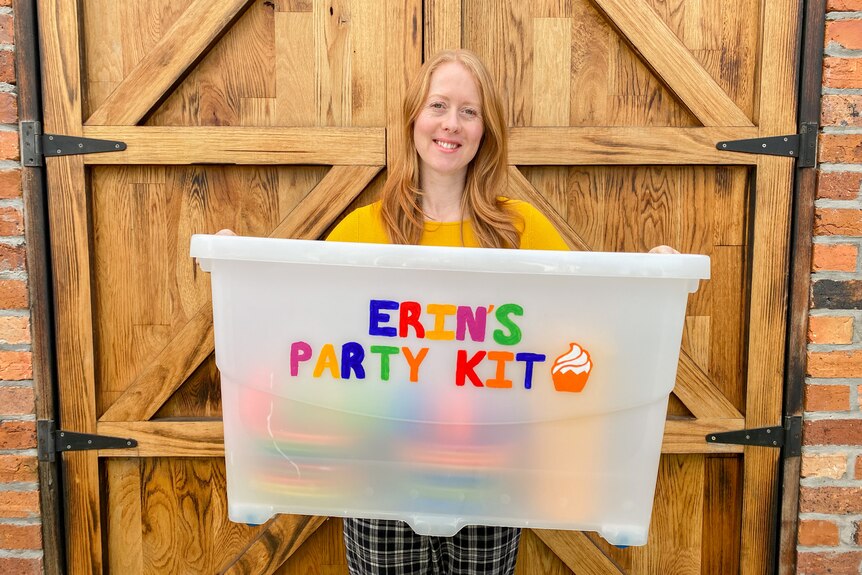A woman holds a large plastic tub labelles 'Erin's Party kit'. 