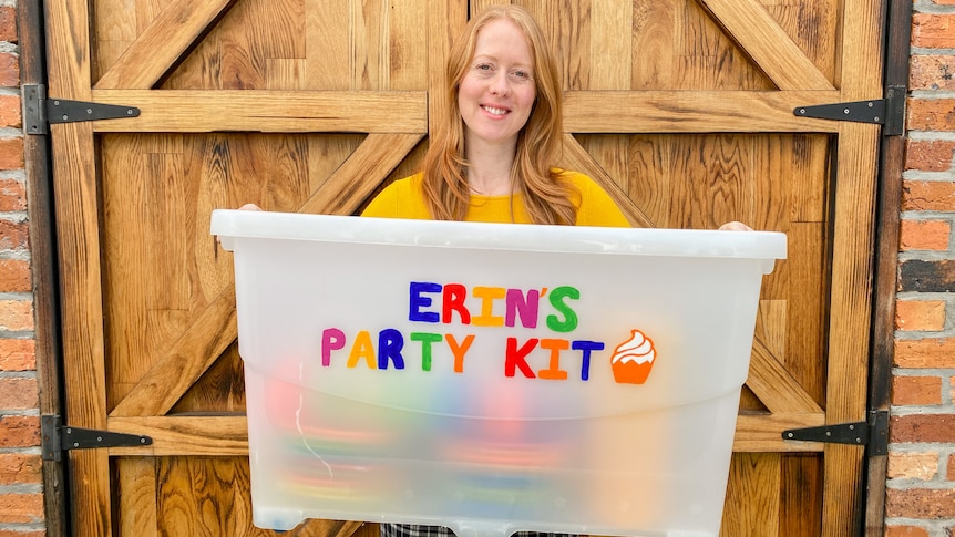 A woman holds a large plastic tub labelles 'Erin's Party kit'. 