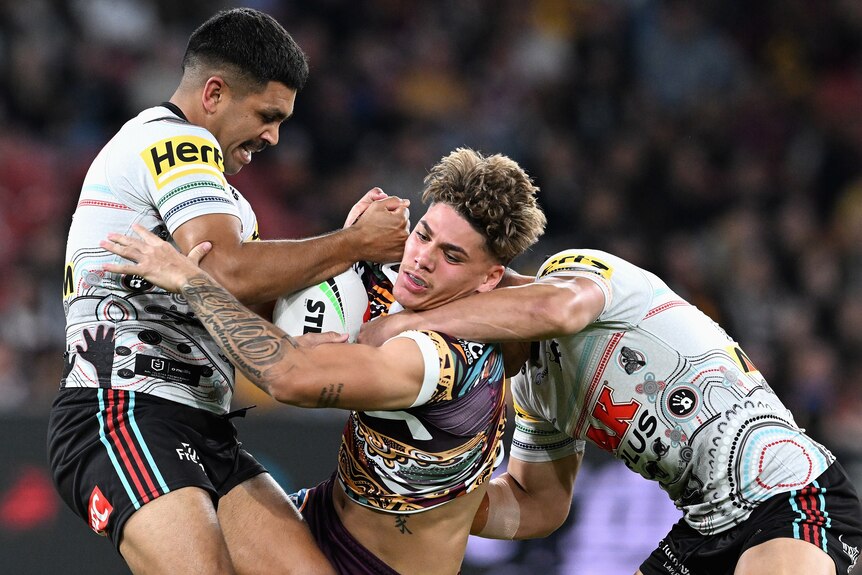 A Brisbane Broncos NRL player holds the ball as he is tackled by two Penrith opponents.