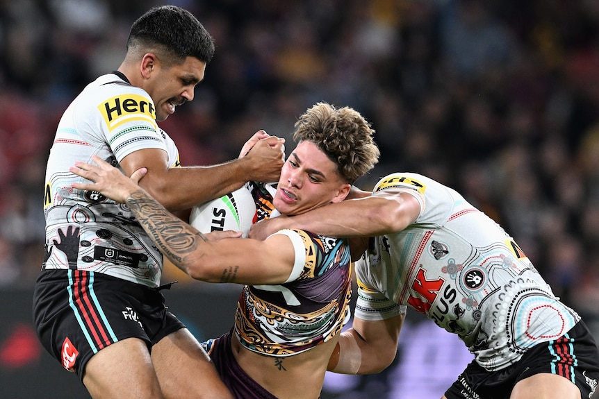 A Brisbane Broncos NRL player holds the ball as he is tackled by two Penrith opponents.