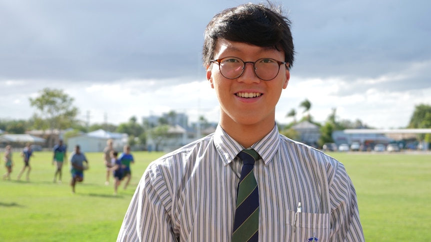 Heetae Lim, shoulders and head shot, smiling at camera, wearing glasses, uniform, grass and kids in background.