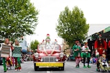 A red car hung with a sign reading "naughty elves" drives down a street lined with people.