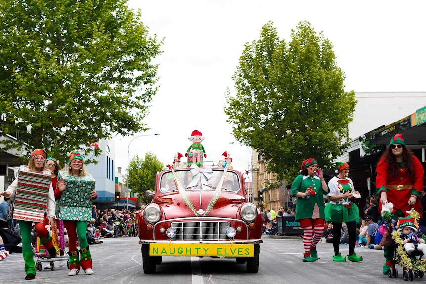 A red car hung with a sign reading "naughty elves" drives down a street lined with people