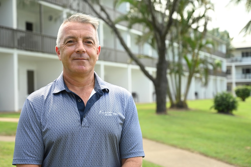 A grey-haired man in uniform smiles at the camera on a clear day with a block of resort units in the background.