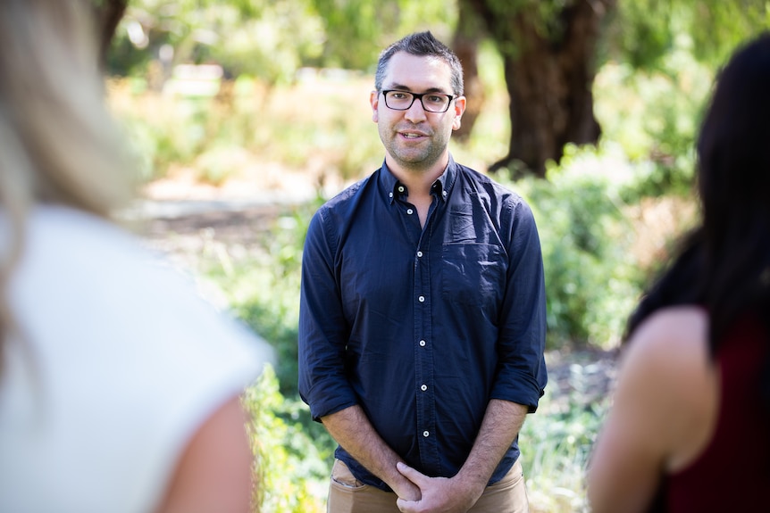 A man in a blue button up shirt with rolled up sleeves talks while standing.