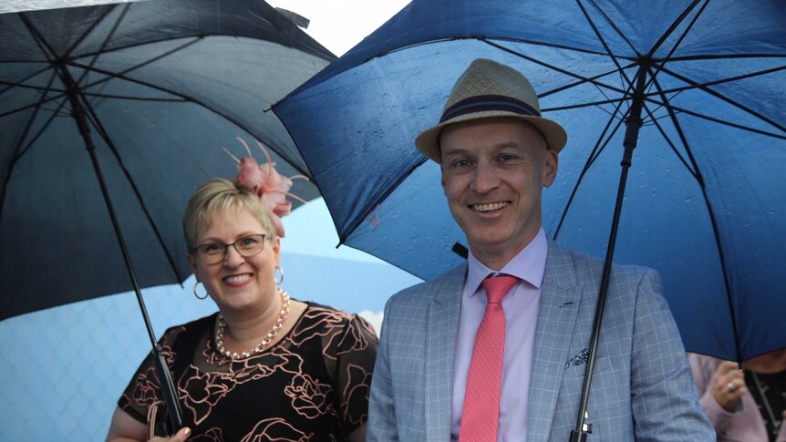 A man in a straw hat and a woman in a fascinator holding umbrellas.