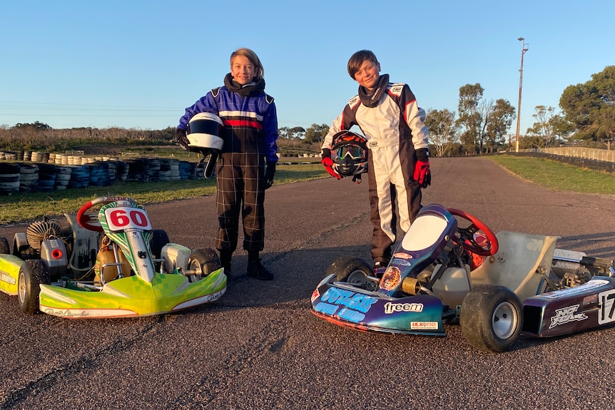Two boys in racing overalls with helmets standing in front of two go-karts on race track lined with tyres