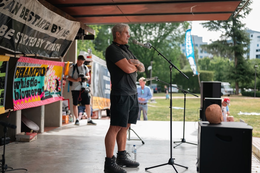 A man stands arms folded in front of a microphone on a small stage. Behind him are banners like 'Champion whistleblowers'.