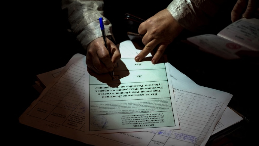 A person signs a ballot paper in a dimly lit room.