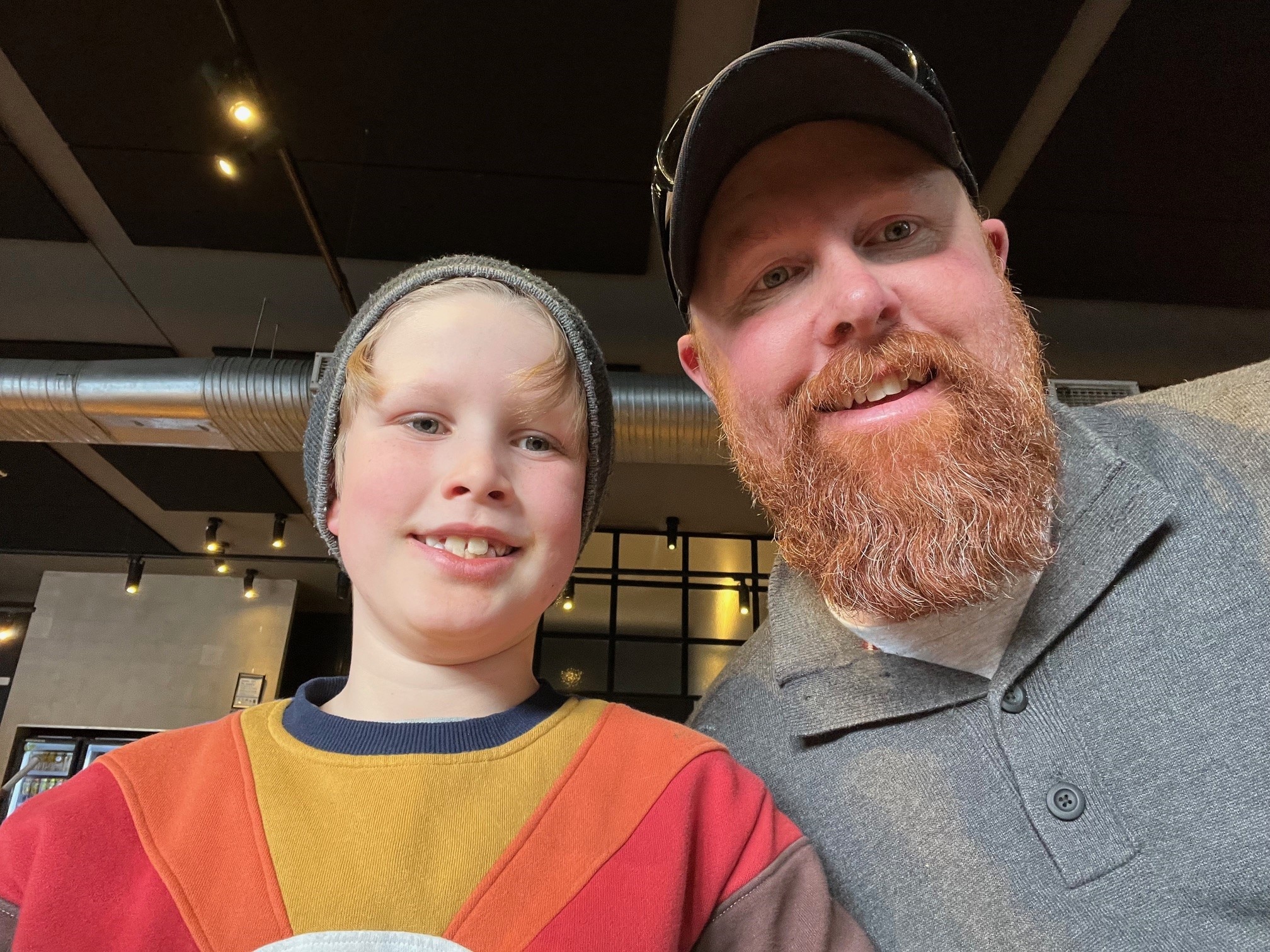 A bearded man with a cap sits with his blonde son, who is wearing a beanie. Both are smiling. 