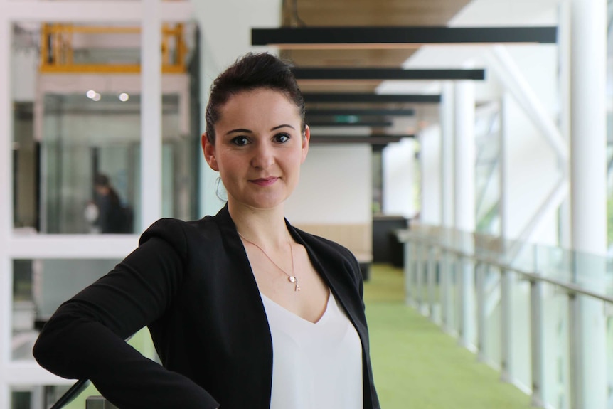 Woman with black jacket, white top and silver necklace stands with arm resting on glass balustrade.