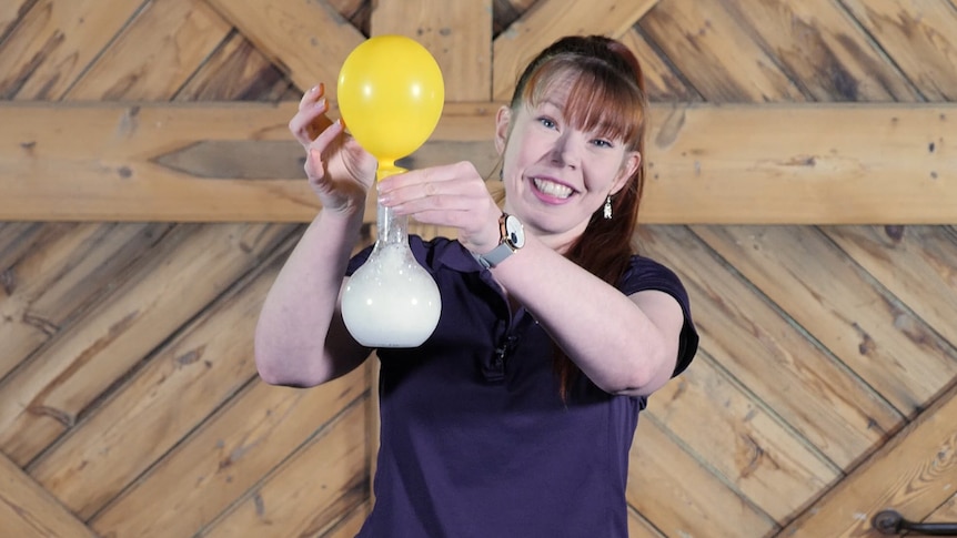 A woman holds a balloon attached to a bottle while doing a science experiment