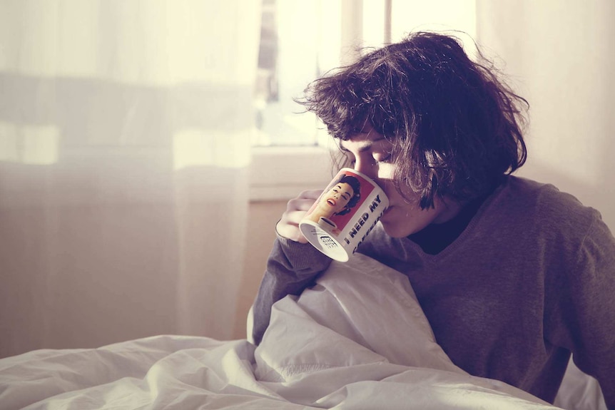 A woman sits in bed with a cup of coffee.