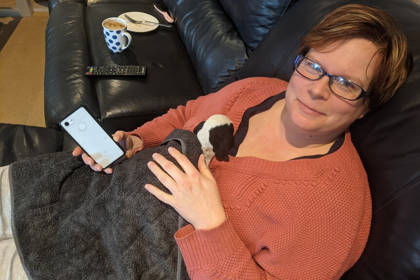A woman reclined on a leather couch with a tiny lamb on her chest, cuddled under a blanket.