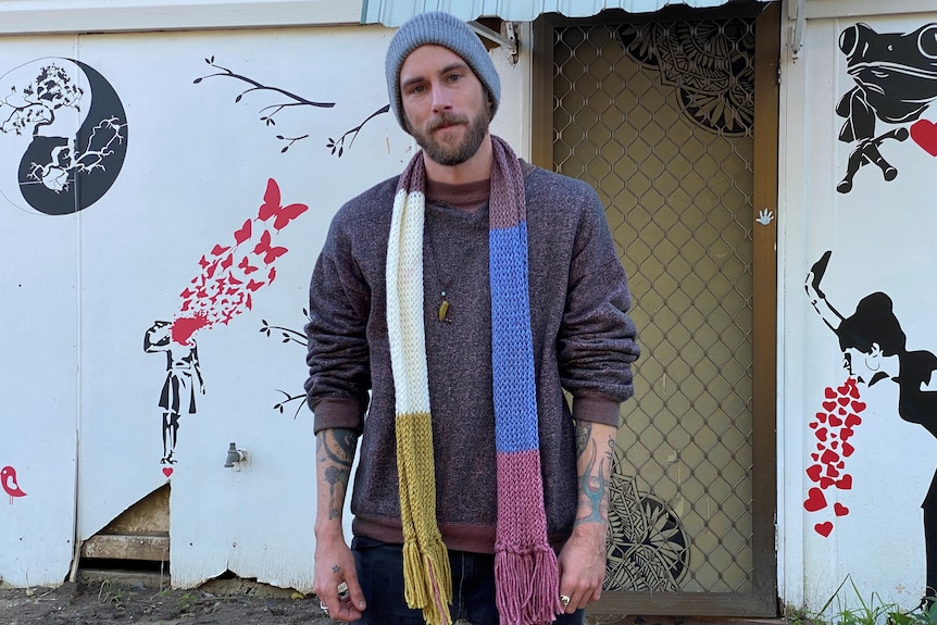 A man wearing a beanie and scarf stands in front of a wall decorated with black and red artwork.