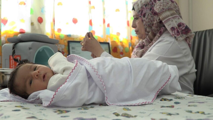 A healthy looking baby wrapped in a white blanket, being treated by a UNRWA doctor who is preparing a vaccine.