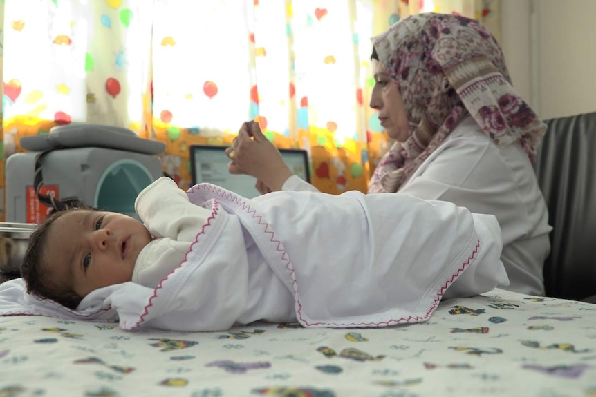 A healthy looking baby wrapped in a white blanket, being treated by a UNRWA doctor who is preparing a vaccine.