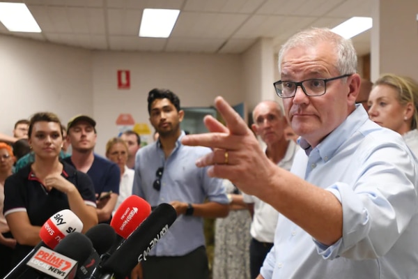 A man in glasses with rolled up sleeves takes questions from a press pack.