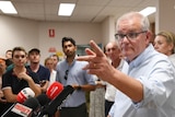A man in glasses with rolled up sleeves takes questions from a press pack.