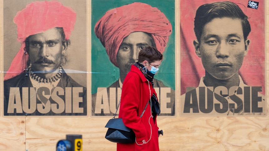 A person with a face mask and red coat walks past a wall with three black and white photos and the word Aussie on it 