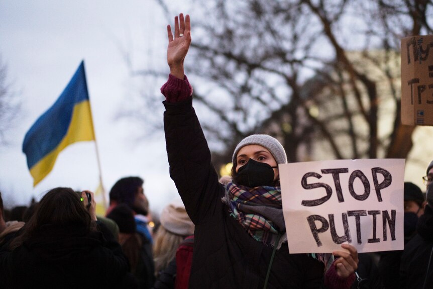 A woman holds a sign that reads "stop Putin", with a Ukrainian flag in the background