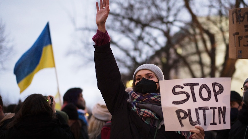 A woman holds a sign that reads stop Putin, with a Ukrainian flag in the background