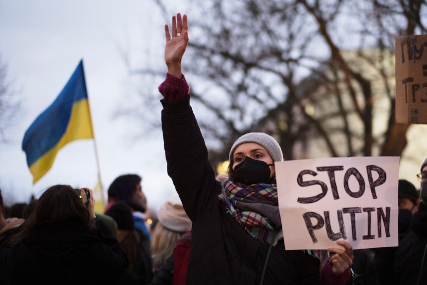 A woman holds a sign that reads "stop Putin", with a Ukrainian flag in the background
