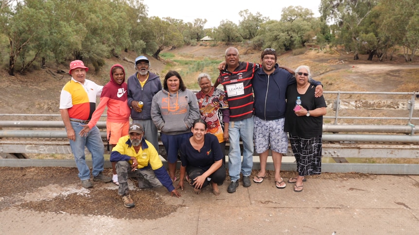 A group of 10 people gathered with arms around each other smiling, with a river bed behind.