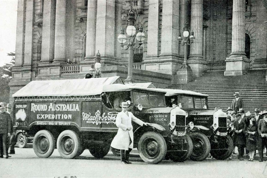 Black and white photo of  a man dressed in white in front of two trucks.