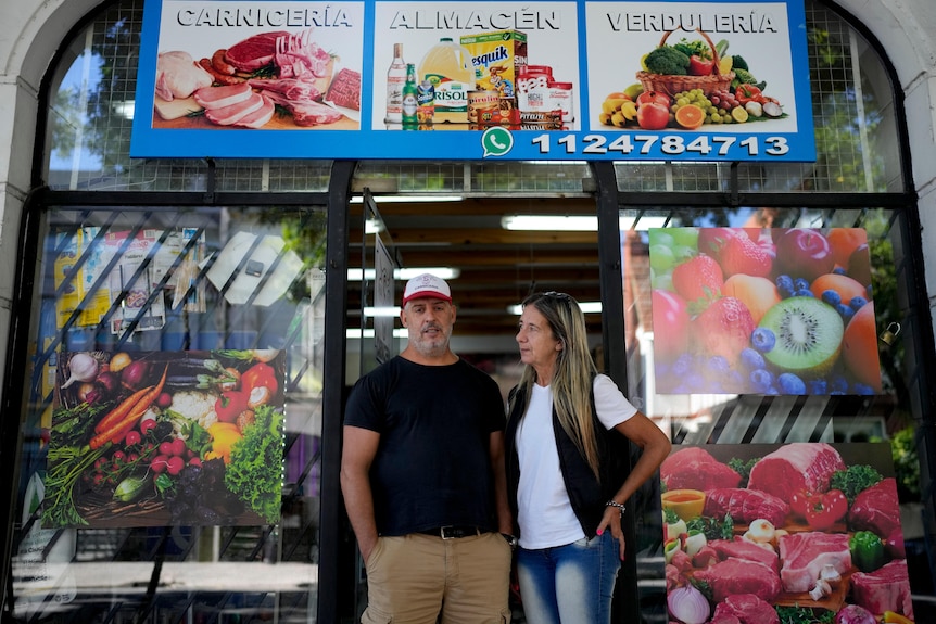 A middle-aged couple stand outside a crowded-looking shop with pictures of vegetables and meat in the window.