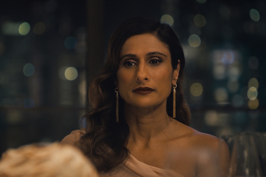 A TV still of Sarayu Blue standing at a party, wearing formalwear. She has a sad expression.