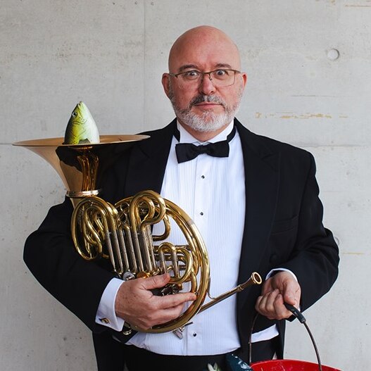 Horn player Peter Luff dressed in black tie, holding a plastic bucket and with a fish coming out of the bell of his horn.