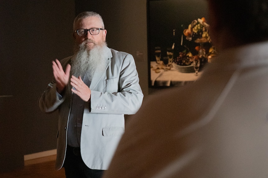 A man with a long white beard and grey jacket in dimly lit art gallery